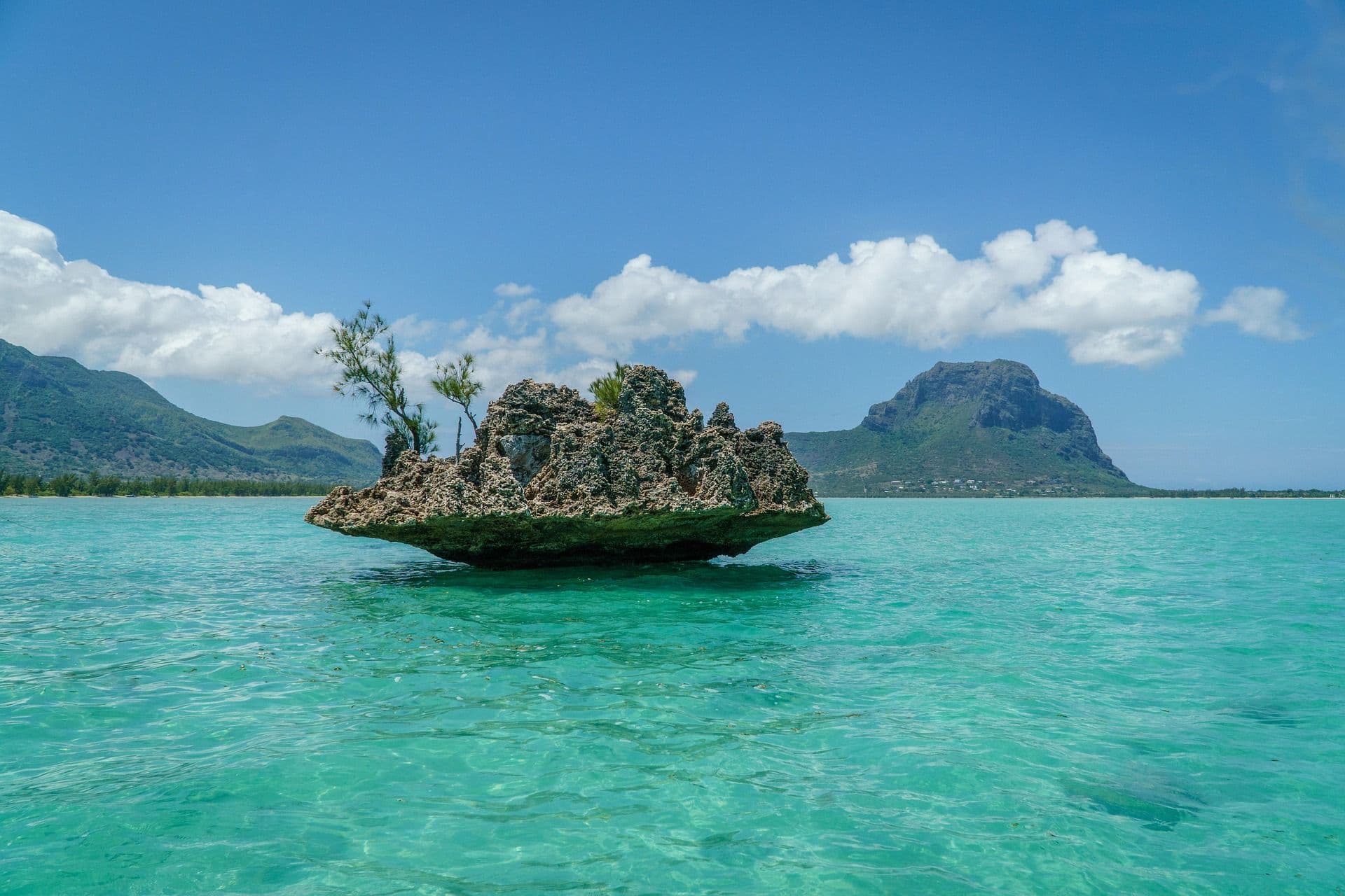 A weathered volcanic rock rising from the turquoise lagoon off Mauritius, with the mountains of the island beyond