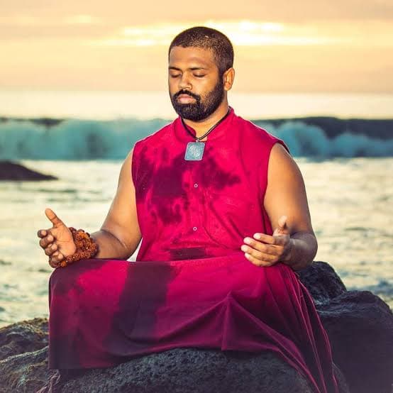 Acharya Dr. Ishan Shivanand Ji seated in meditation on the rocks by the ocean at sunset